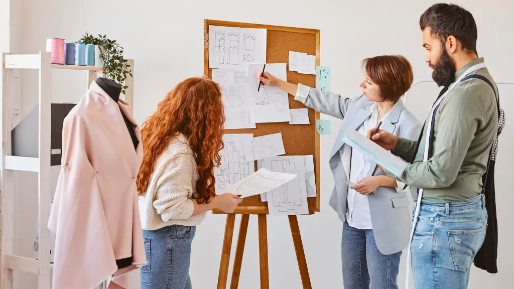 Duas mulheres e um homem em uma loja de roupas analisando designs e o sistema de gestão para loja de roupas em um quadro de cortiça.